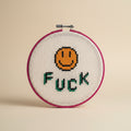 A circular embroidery hoop holding a cream-colored cross-stitch canvas with a stitched orange smiley face and the word 'FUCK' in green and black letters. A hand is holding the hoop against a plain light gray wall.