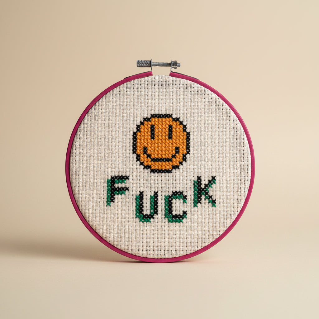 A circular embroidery hoop holding a cream-colored cross-stitch canvas with a stitched orange smiley face and the word 'FUCK' in green and black letters. A hand is holding the hoop against a plain light gray wall.