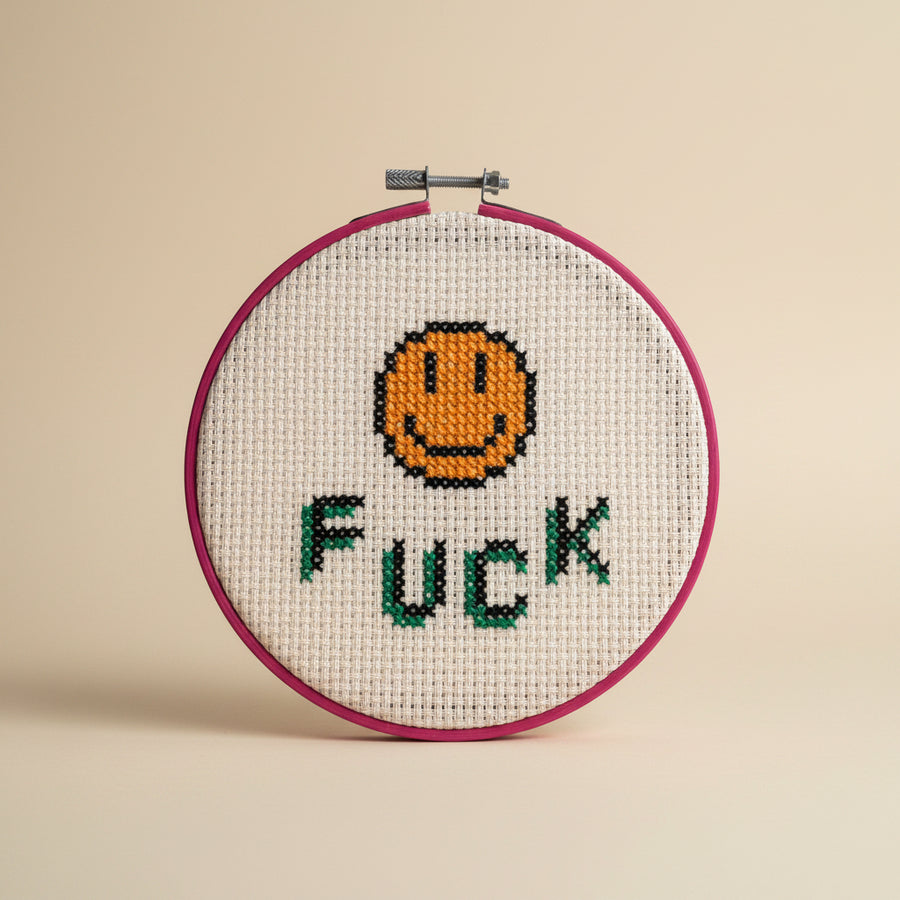 A circular embroidery hoop holding a cream-colored cross-stitch canvas with a stitched orange smiley face and the word 'FUCK' in green and black letters. A hand is holding the hoop against a plain light gray wall.
