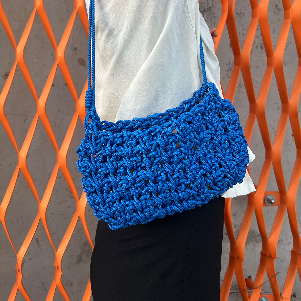 A person wearing a white shirt and black pants carries a large, chunky crocheted blue shoulder bag with a matching blue strap, standing in front of an orange metal grid fence.