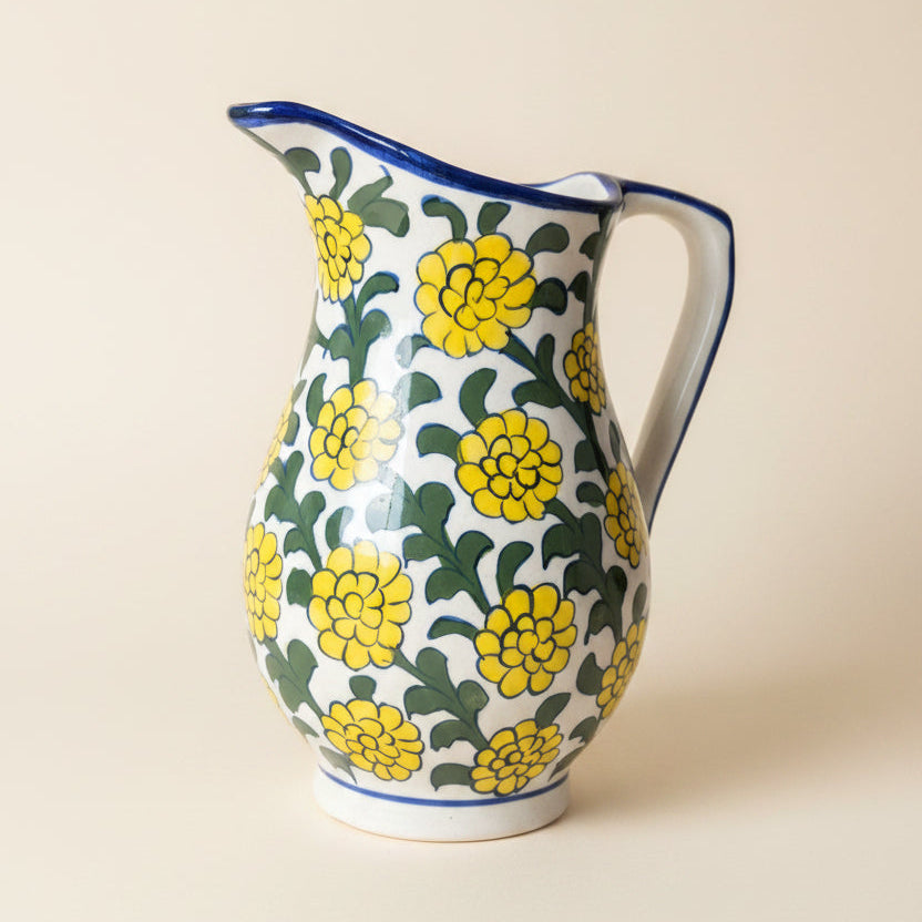 A white ceramic pitcher decorated with a yellow floral pattern and green leaves, featuring a blue rim and handle