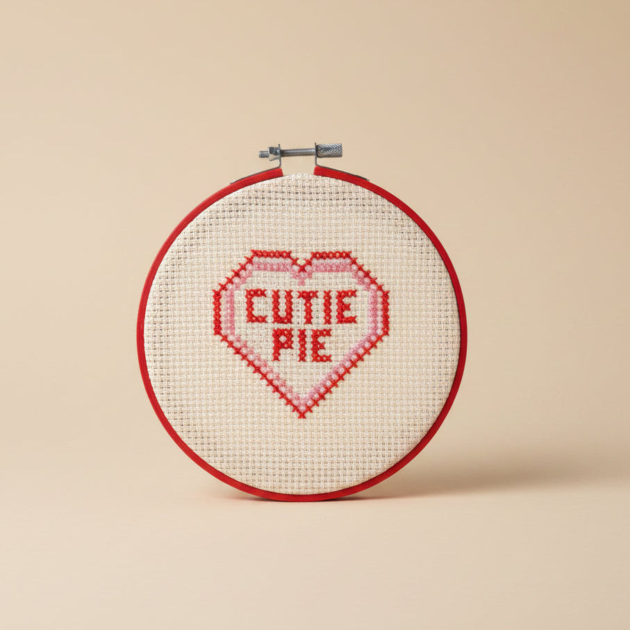 A circular embroidery hoop with cream-colored cross-stitch fabric featuring a red heart outline and the words 'CUTIE PIE' stitched inside in red and pink.
