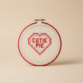 A circular embroidery hoop with cream-colored cross-stitch fabric featuring a red heart outline and the words 'CUTIE PIE' stitched inside in red and pink.